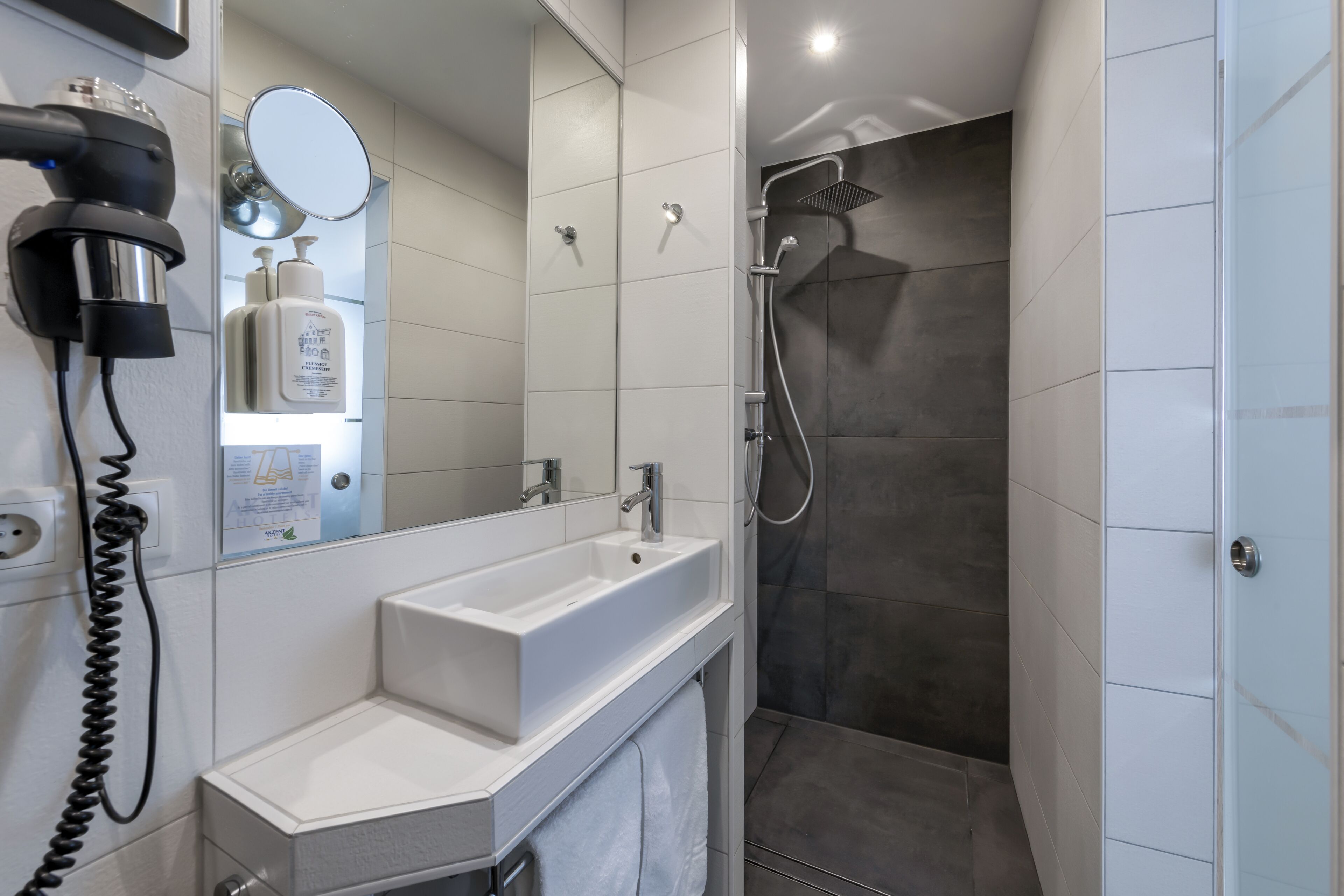 A modern bathroom with a white sink, a mirror, a hairdryer, and a shower.