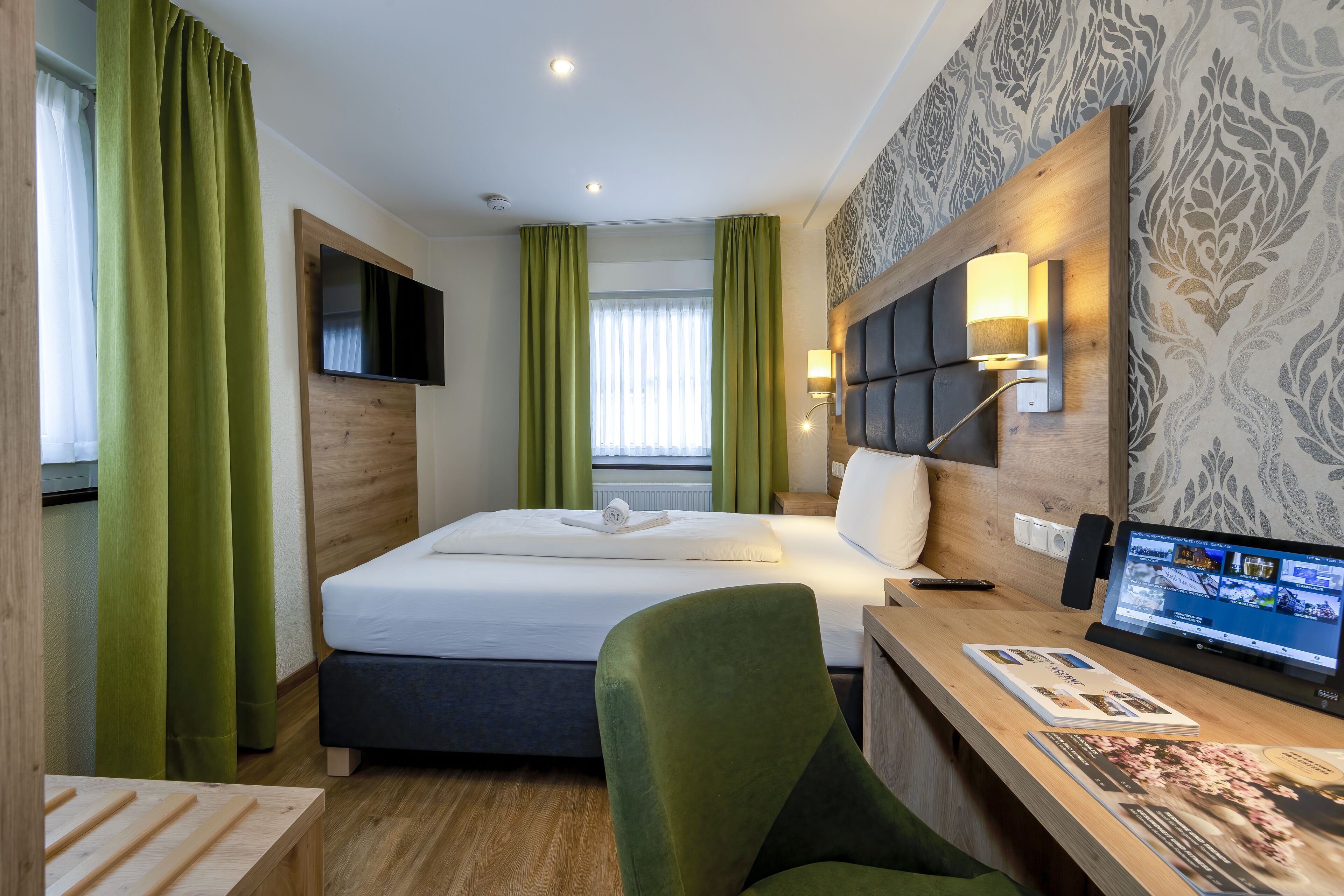 A hotel room with a bed, a desk with a computer, a TV, and green curtains.