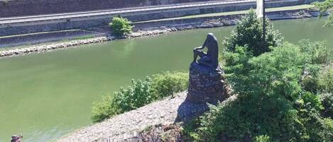 A statue on a stone platform overlooking a river, with a flagpole and greenery in the foreground.