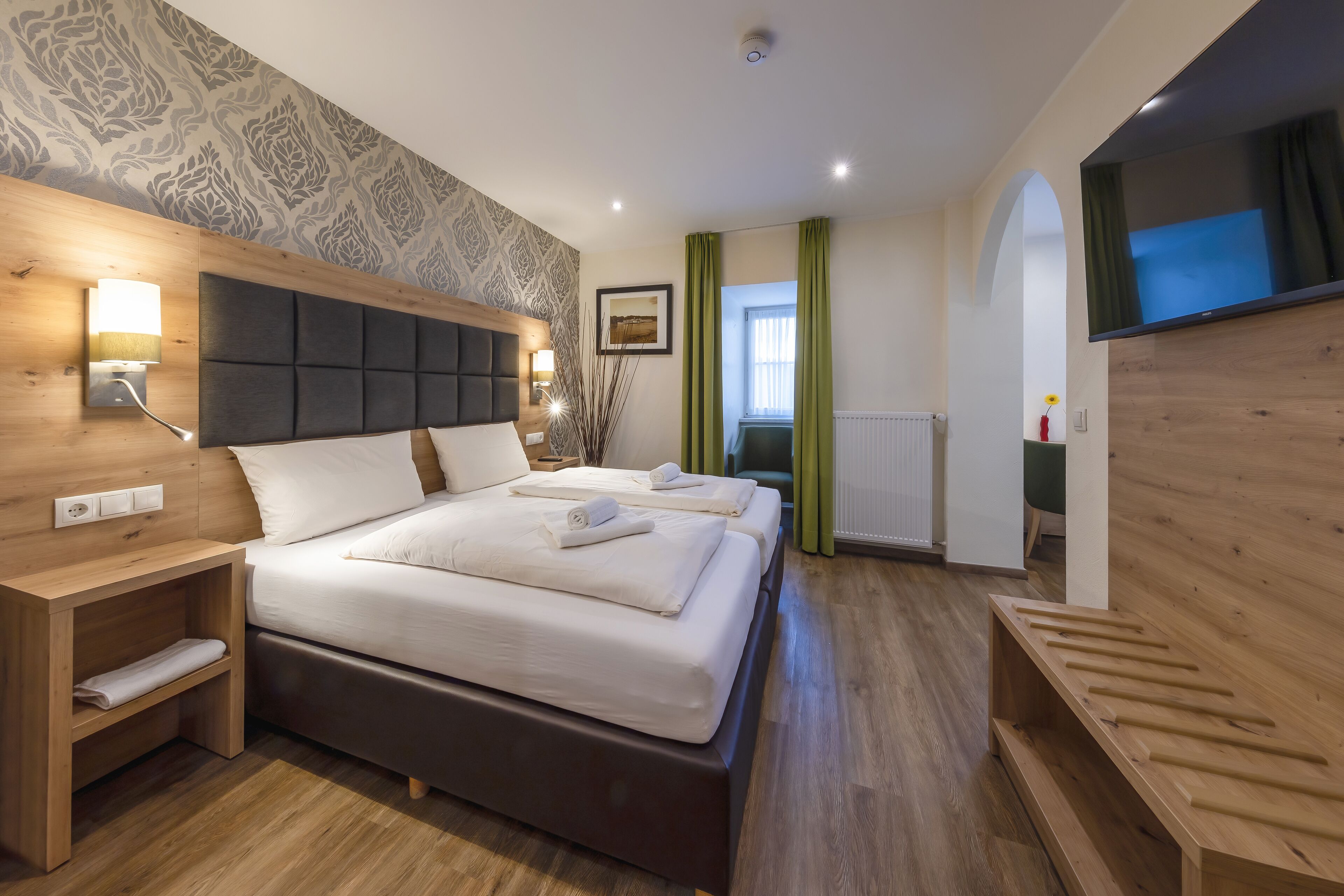 A modern hotel room with two beds, wooden headboards, a large TV, and a wooden bench.