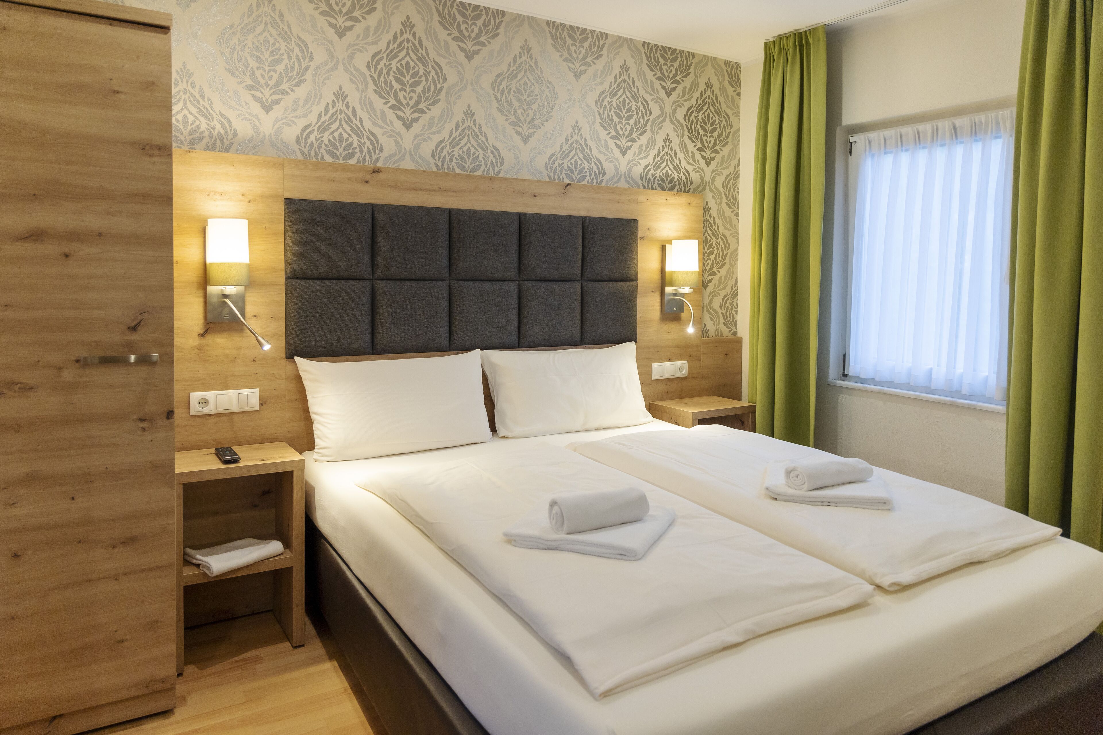 A double bed with white linens, a wooden headboard, bedside lamps, and a window with green curtains.