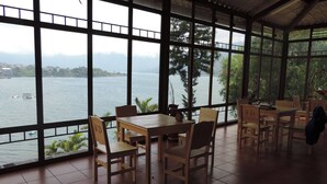 A covered outdoor seating area with wooden tables and chairs, large windows offering a view of a lake and distant mountains.