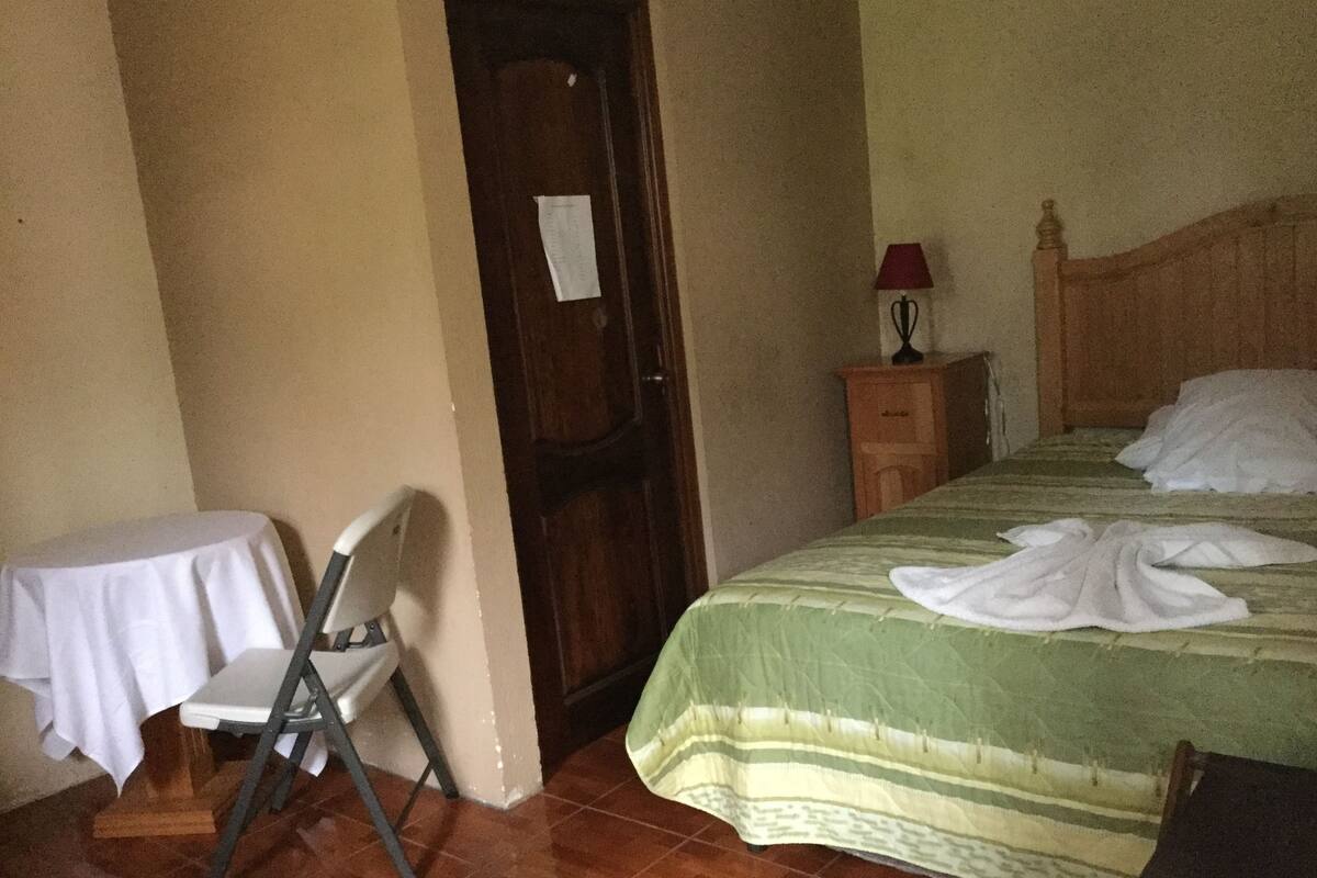 A room with a bed, a chair, a table, a nightstand, and a door.