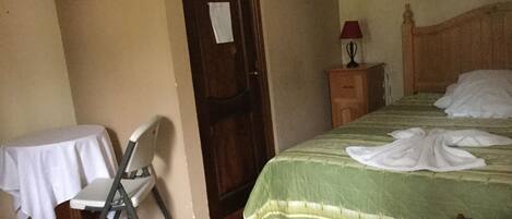Um quarto com cama, uma cadeira, uma mesa, mesa de cabeceira e uma porta.