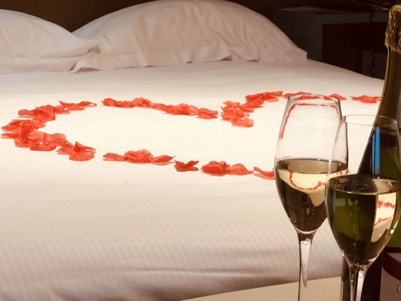 Una cama con pétalos de rosa dispuestos en forma de corazón y dos copas de vino blanco sobre la mesita de noche.