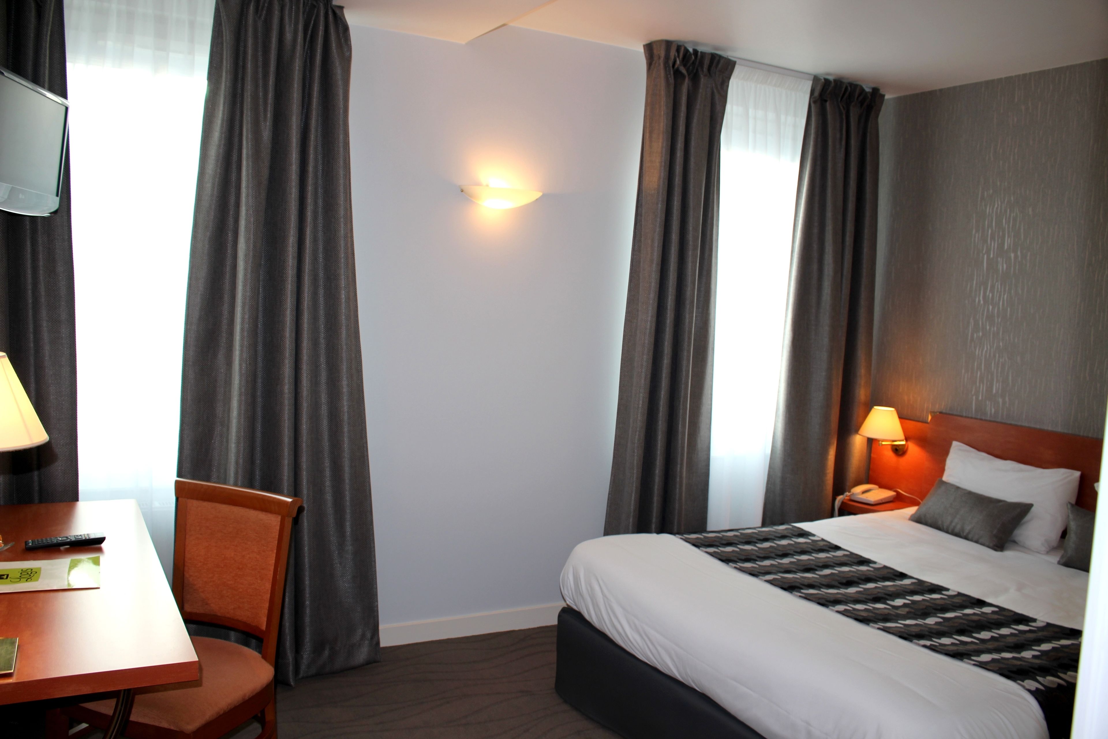 Habitación de hotel con cama, escritorio con silla, lámpara y mesita de noche con teléfono.