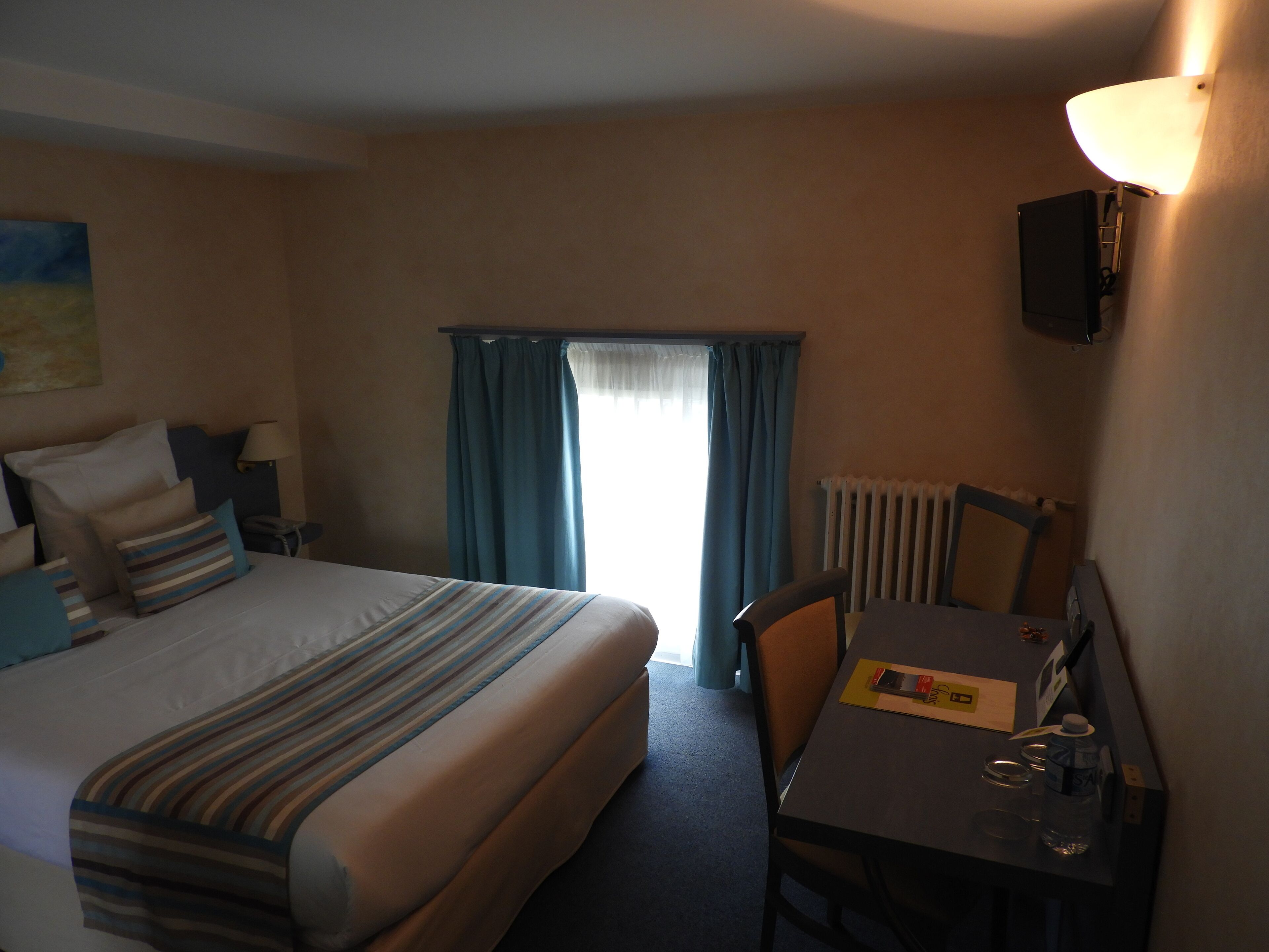 Quarto de hotel com cama, mesa de cabeceira, luminária, poltrona e escrivaninha com cadeira.