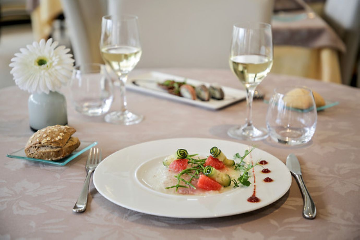 Una mesa de comedor con un plato de comida blanca, dos copas de vino blanco, un pequeño jarrón con una flor blanca y un vaso de agua.