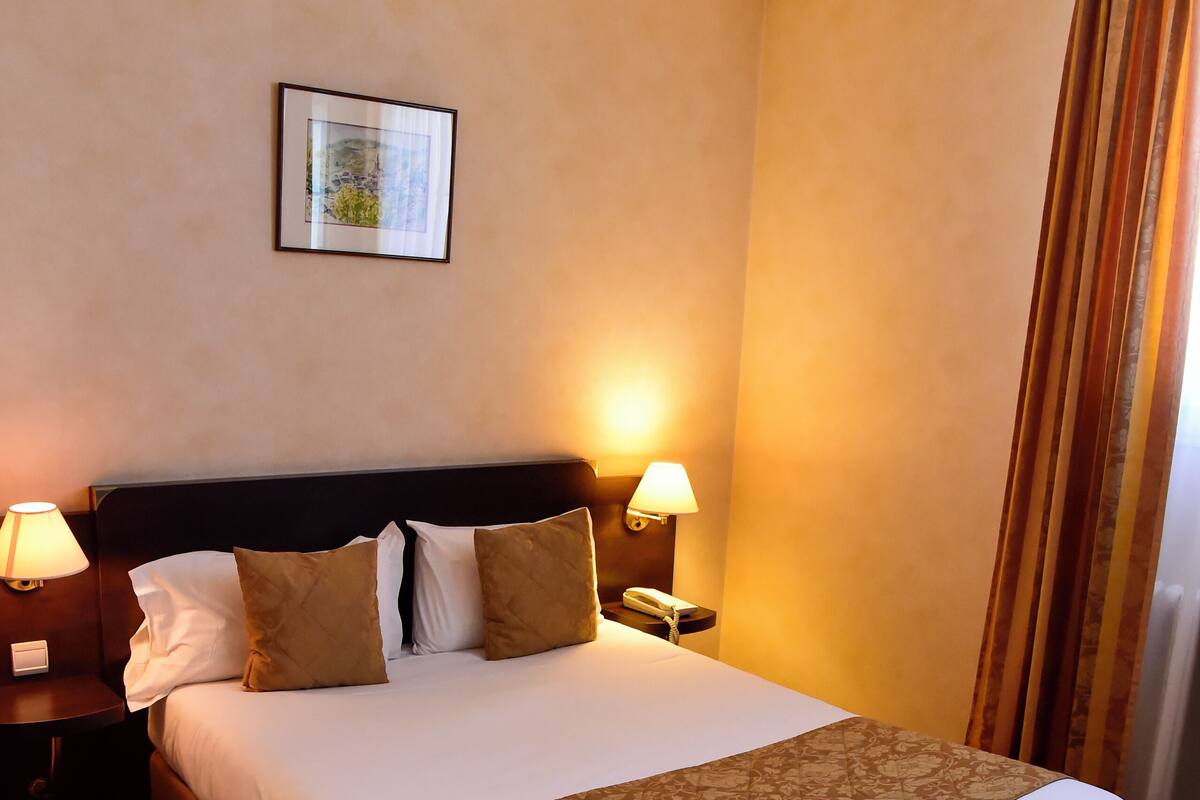 Ein Hotelzimmer mit einem großen Bett, zwei Nachttischlampen, einem Nachttisch, einem gerahmten Bild an der Wand und einem Fenster mit Vorhängen.