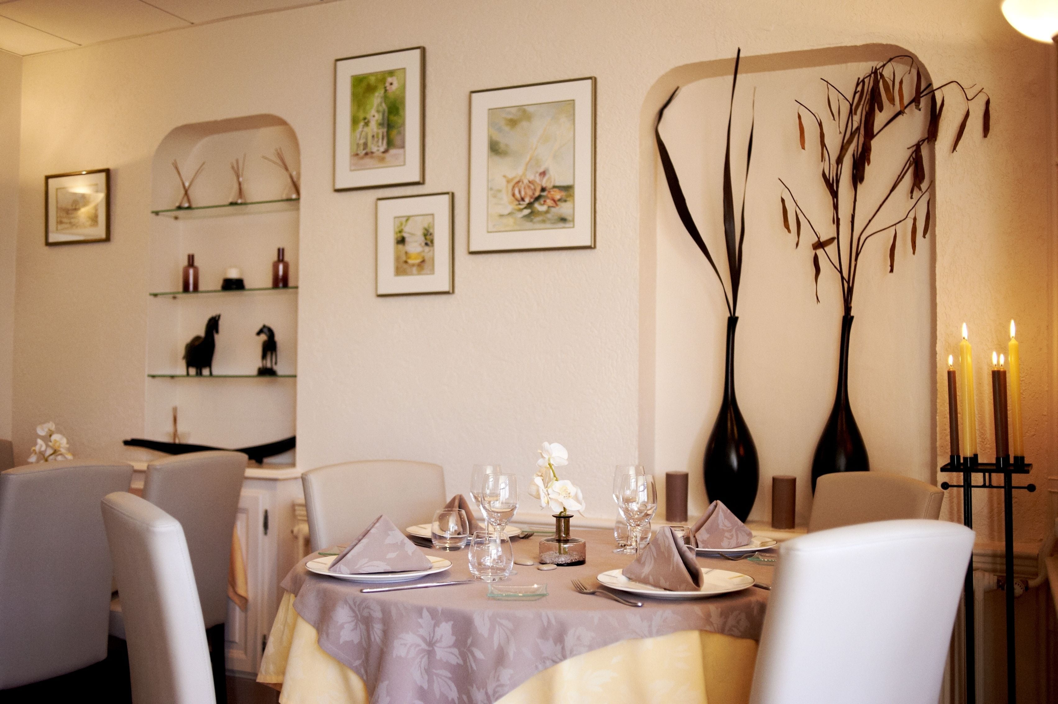 A dining area with a table set for four, surrounded by chairs. There are framed artworks on the wall, a vase with dried branches, and a candle holder with candles.