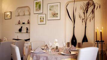 A dining area with a table set for four, surrounded by chairs. There are framed artworks on the wall, a vase with dried branches, and a candle holder with candles.