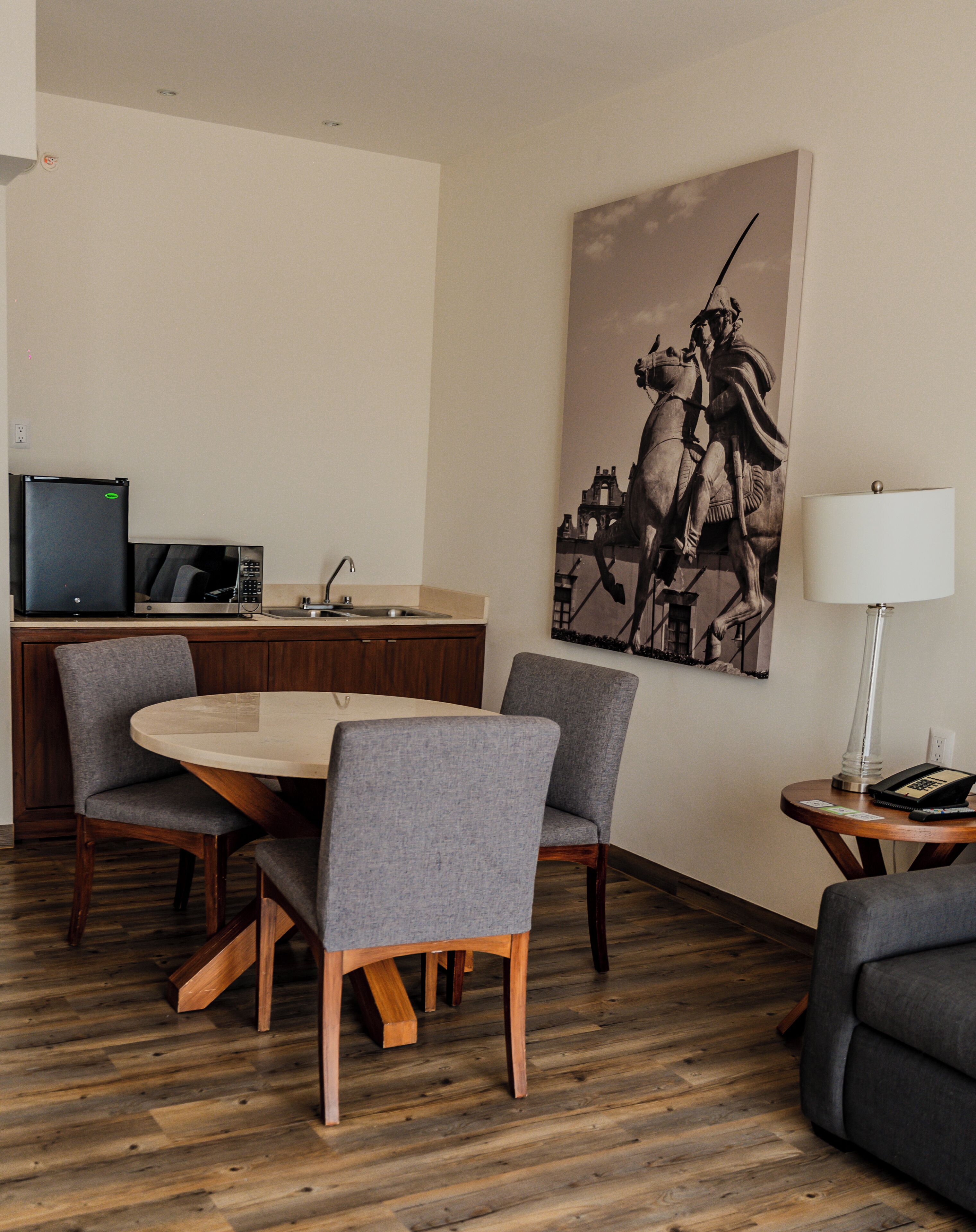 Habitación de hotel moderna con una mesa de comedor, sillas, una zona de cocina con microondas y refrigerador, un cuadro grande y una lámpara.