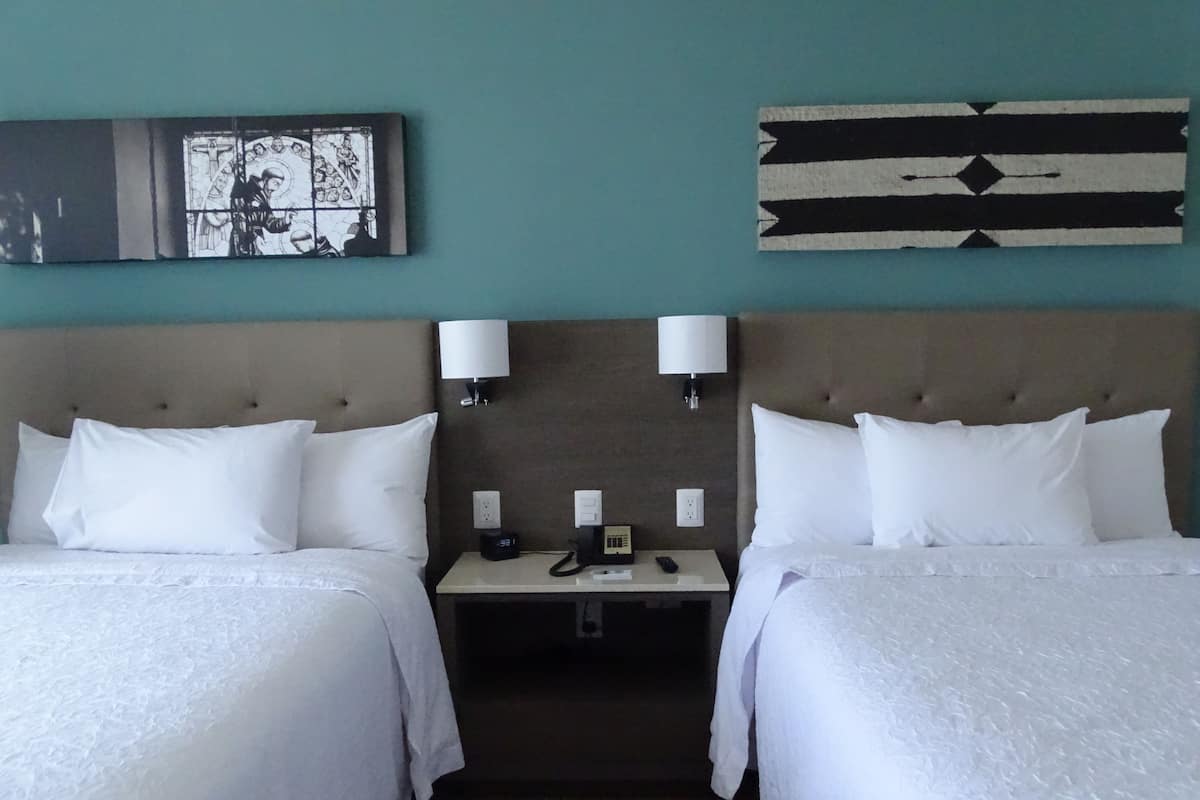 Habitación de hotel con dos camas, una mesita de noche con teléfono y lámpara, y una pared decorada con un cuadro enmarco y una obra de arte geométrica.