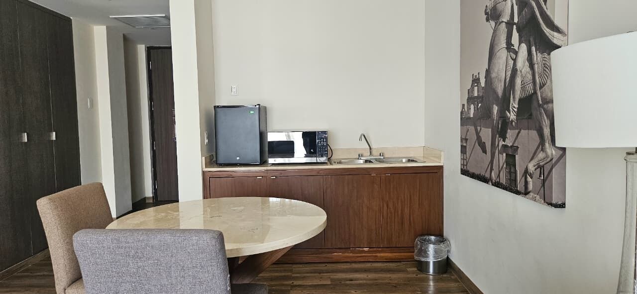 A compact kitchen with a microwave, sink, and a small refrigerator. There is a round table with two chairs and a large painting on the wall.