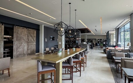 A spacious hotel lobby with a long communal table, seating areas, and large windows.