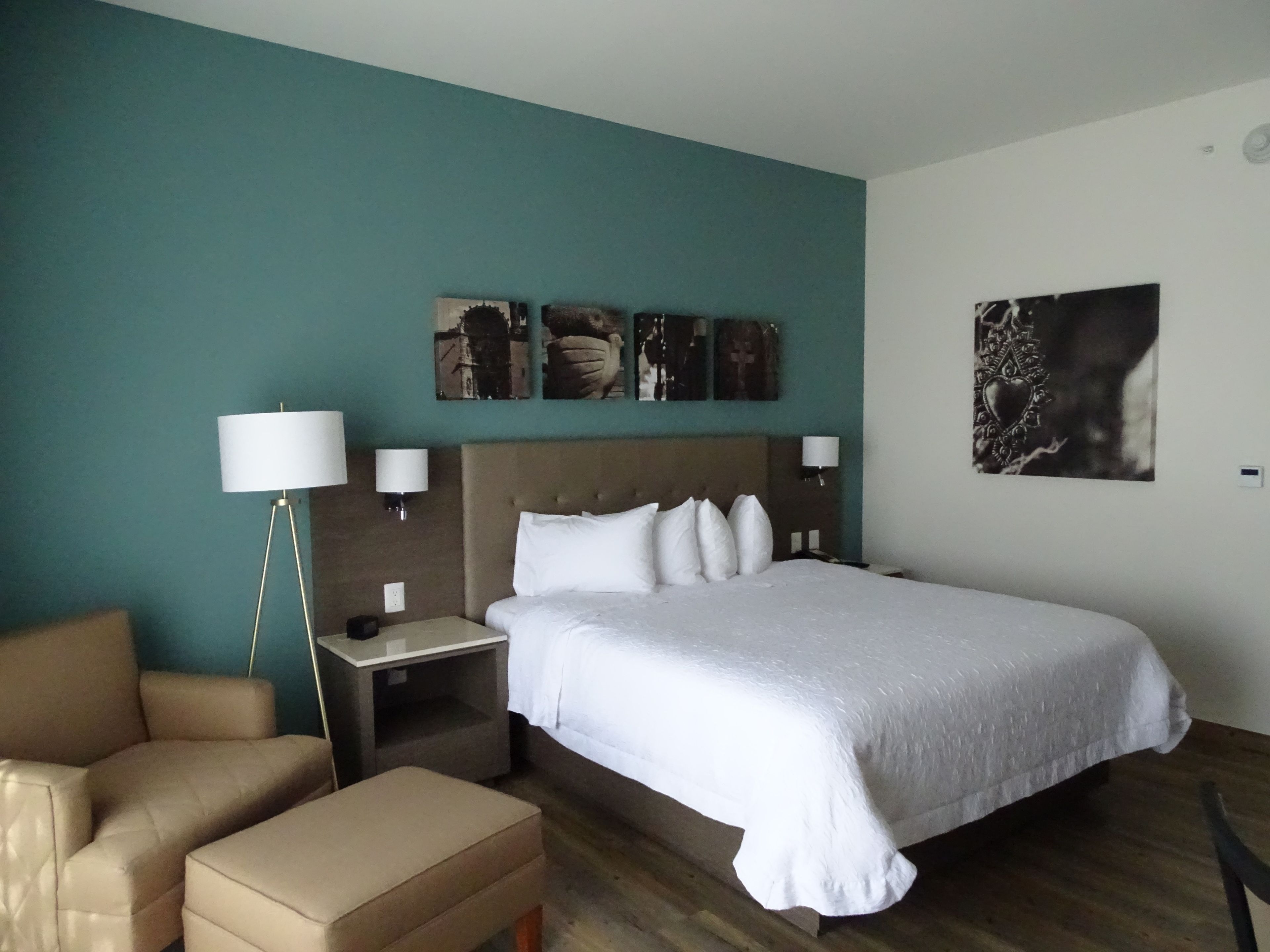 A hotel room with a large bed, a nightstand, a lamp, a chair, and a wall with artwork.