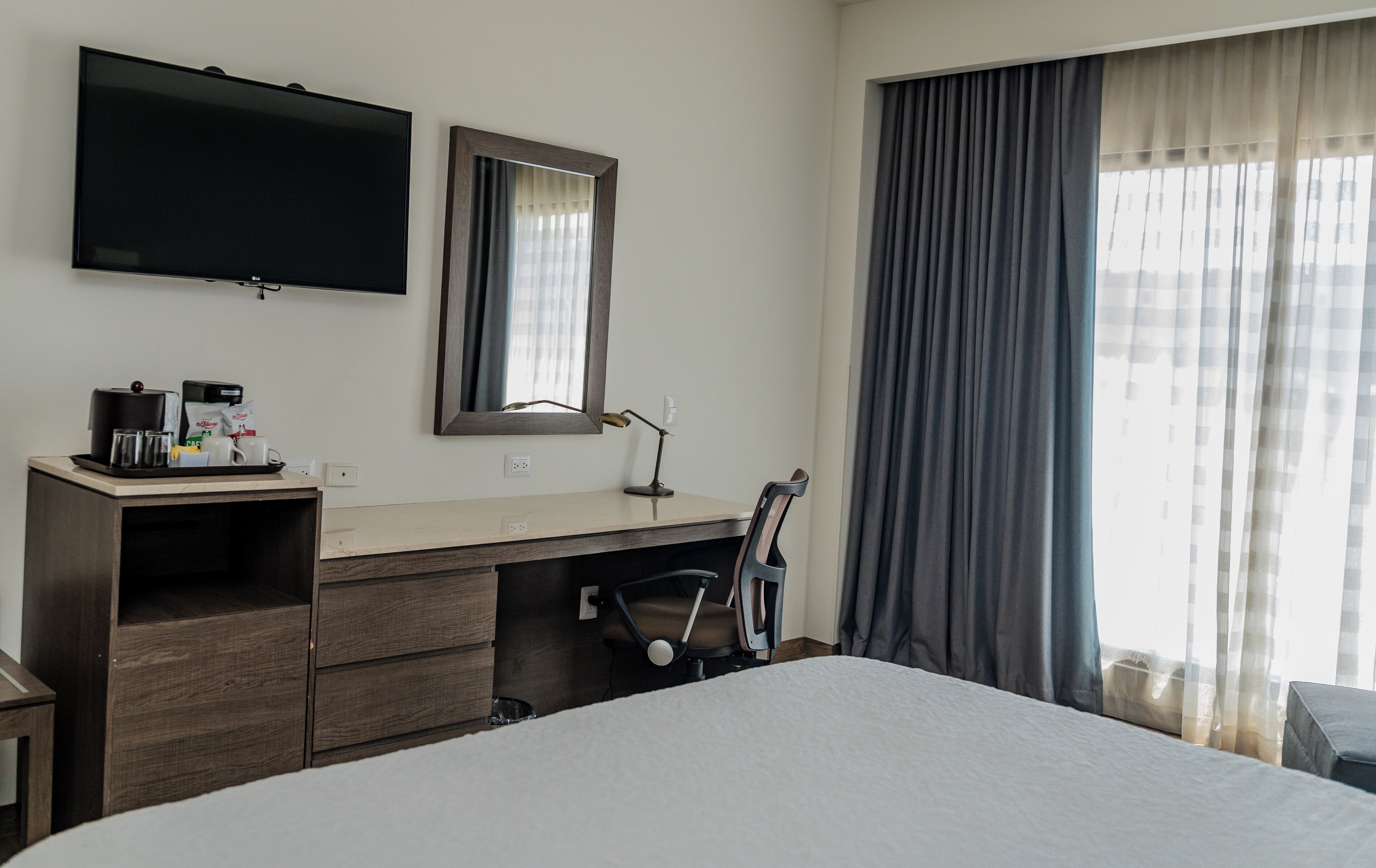 A hotel room with a bed, a desk with a computer, a TV, and a window with curtains.