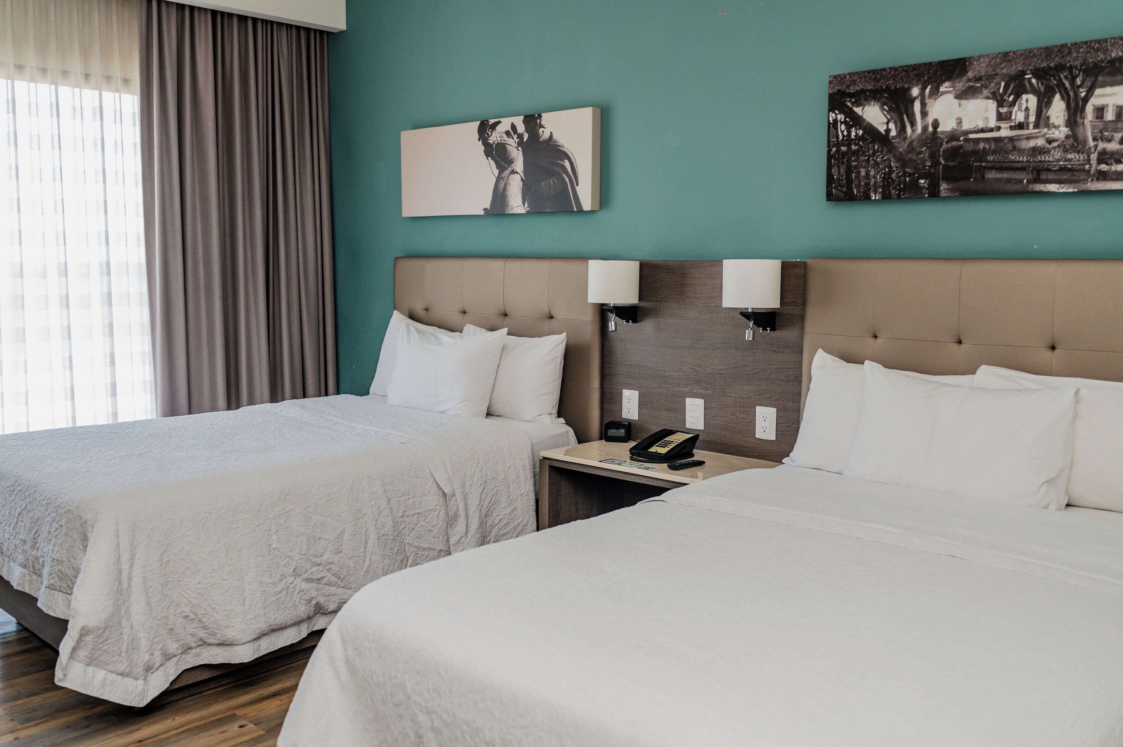 Habitación de hotel con dos camas, una mesita de noche con teléfono, decoración en la pared y una ventana con cortinas.