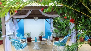 A covered outdoor seating area with white wicker chairs and a small round table, surrounded by potted plants and draped fabric. - Hotel Tronco Inc (Boca Chica)