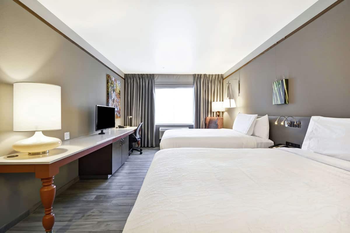 A hotel room with two beds, a desk, a TV, and a window with curtains.