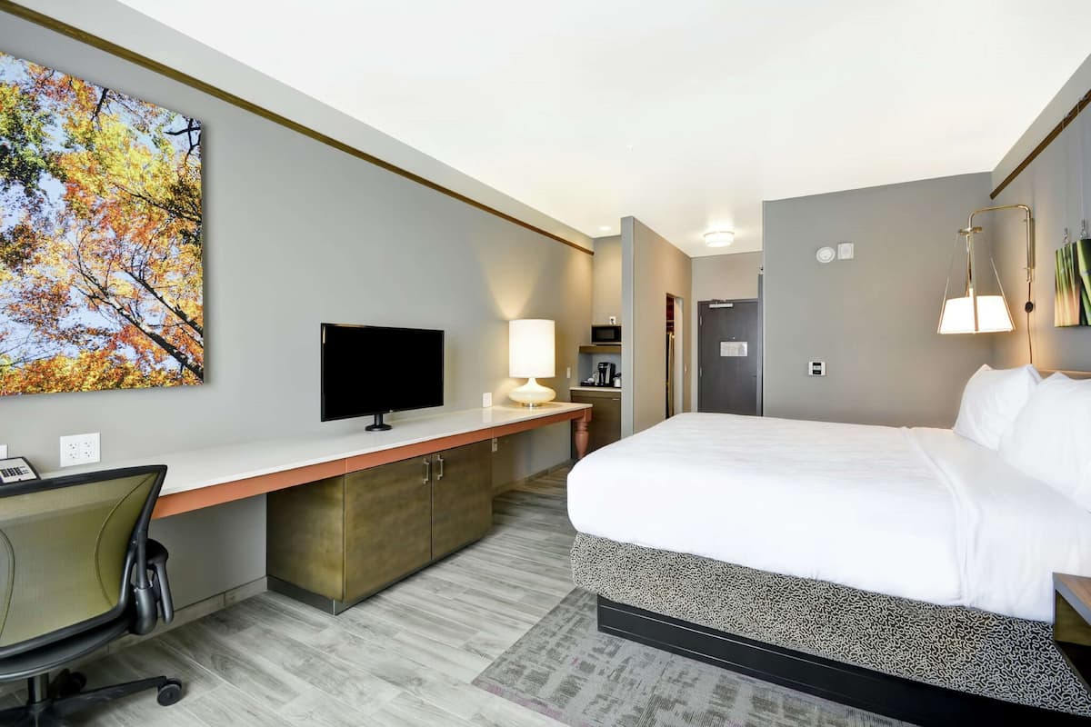 A hotel room with a large bed, a desk with a computer, a TV, and a painting of autumn leaves on the wall.
