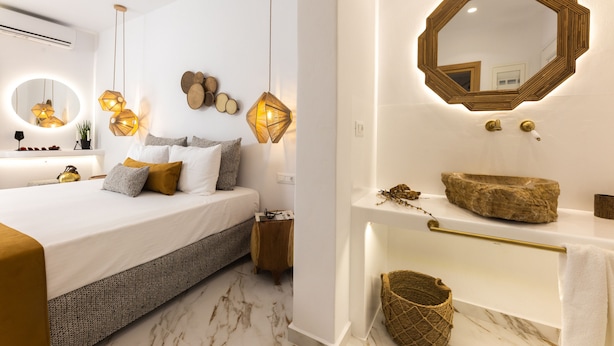 A modern bedroom with a large bed, bedside tables, a round mirror, and a bathroom with a vanity and sink. - Ktima Lino (Naxos)