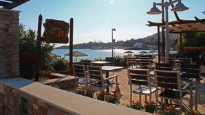 An outdoor seating area with tables and chairs, a sign for Olympia Tavern, and a view of the sea and cityscape. - Olympia Hotel (Syros)