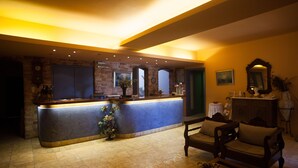 A reception area with a stone wall, a reception desk, a clock, and a seating area with a chair and a small table. - Olympia Hotel (Syros)