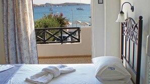 A hotel room with a bed, a wooden nightstand, a window with a view of the sea and boats, and a balcony with a railing. - Olympia Hotel (Syros)