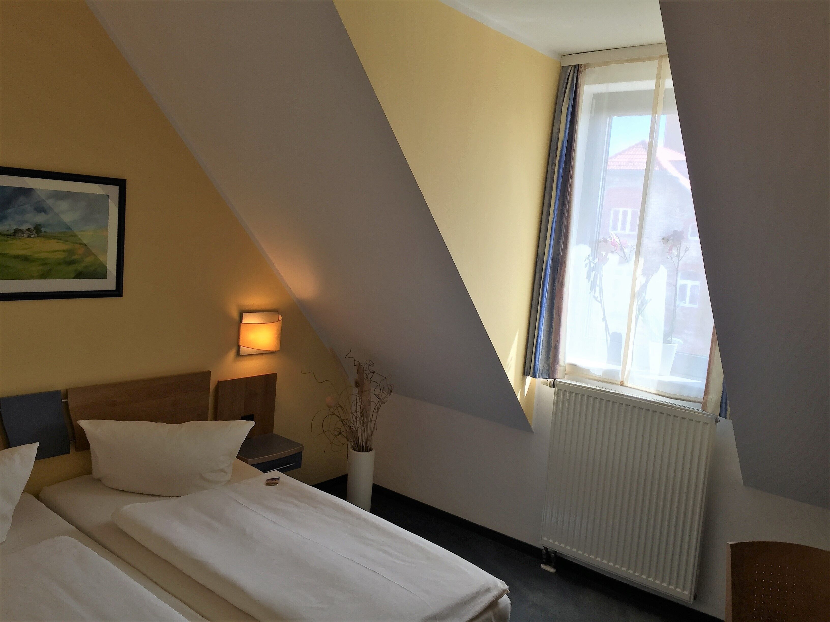Ein Schlafzimmer mit einem Bett, einem Nachttisch, einer Lampe und einem Fenster mit Blick auf Gebäude.