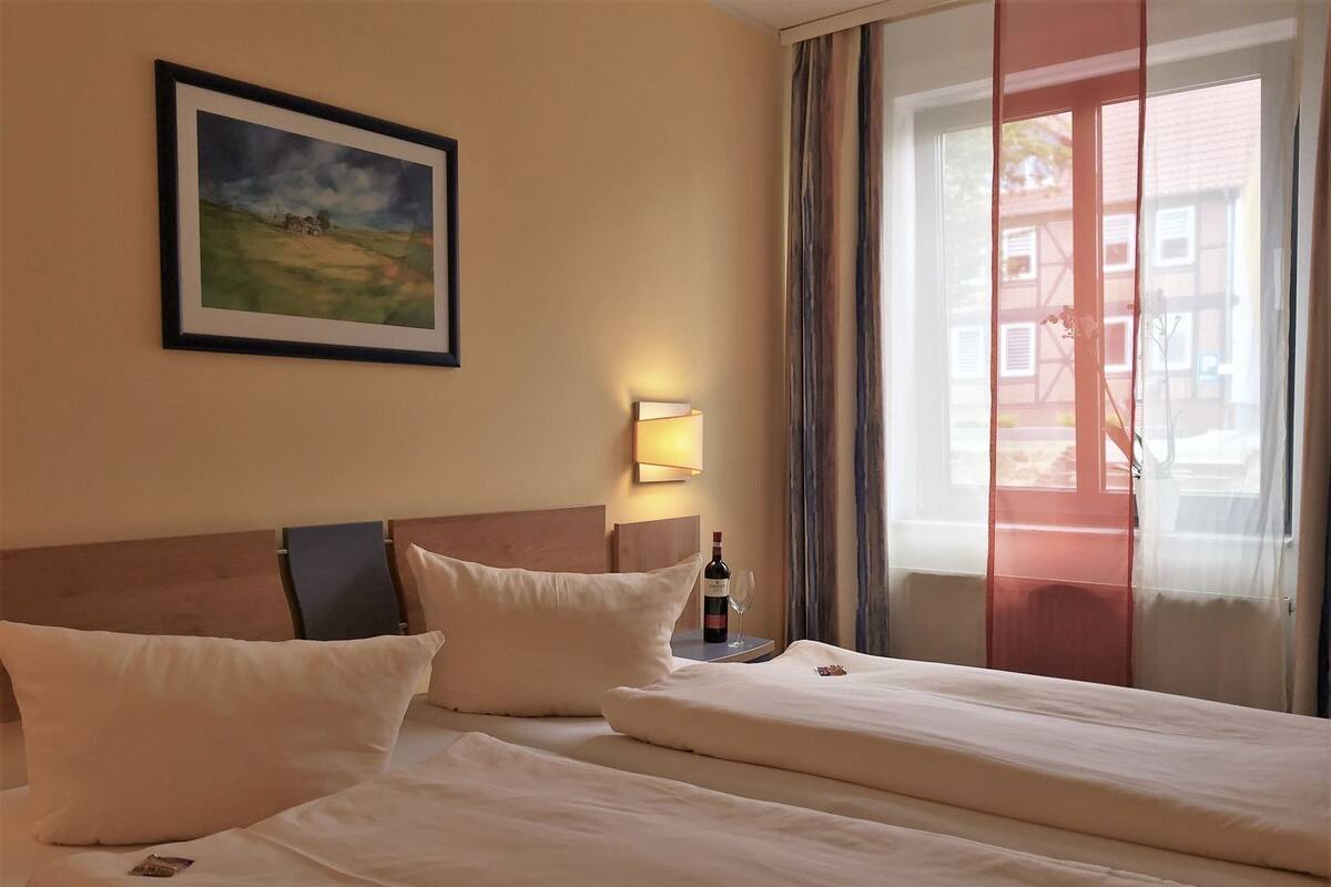 Une chambre d’hôtel avec deux lits, un tableau encadré au mur, une bouteille de vin et une fenêtre avec des rideaux.