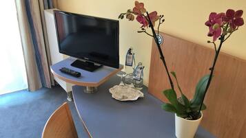 A hotel room with a television on a stand, a remote control, a small table with a glass of water, a bottle of hand sanitizer, and a potted orchid plant.