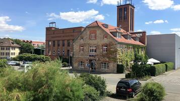 A historic building with a stone facade and a red-tiled roof, surrounded by greenery and parked cars.