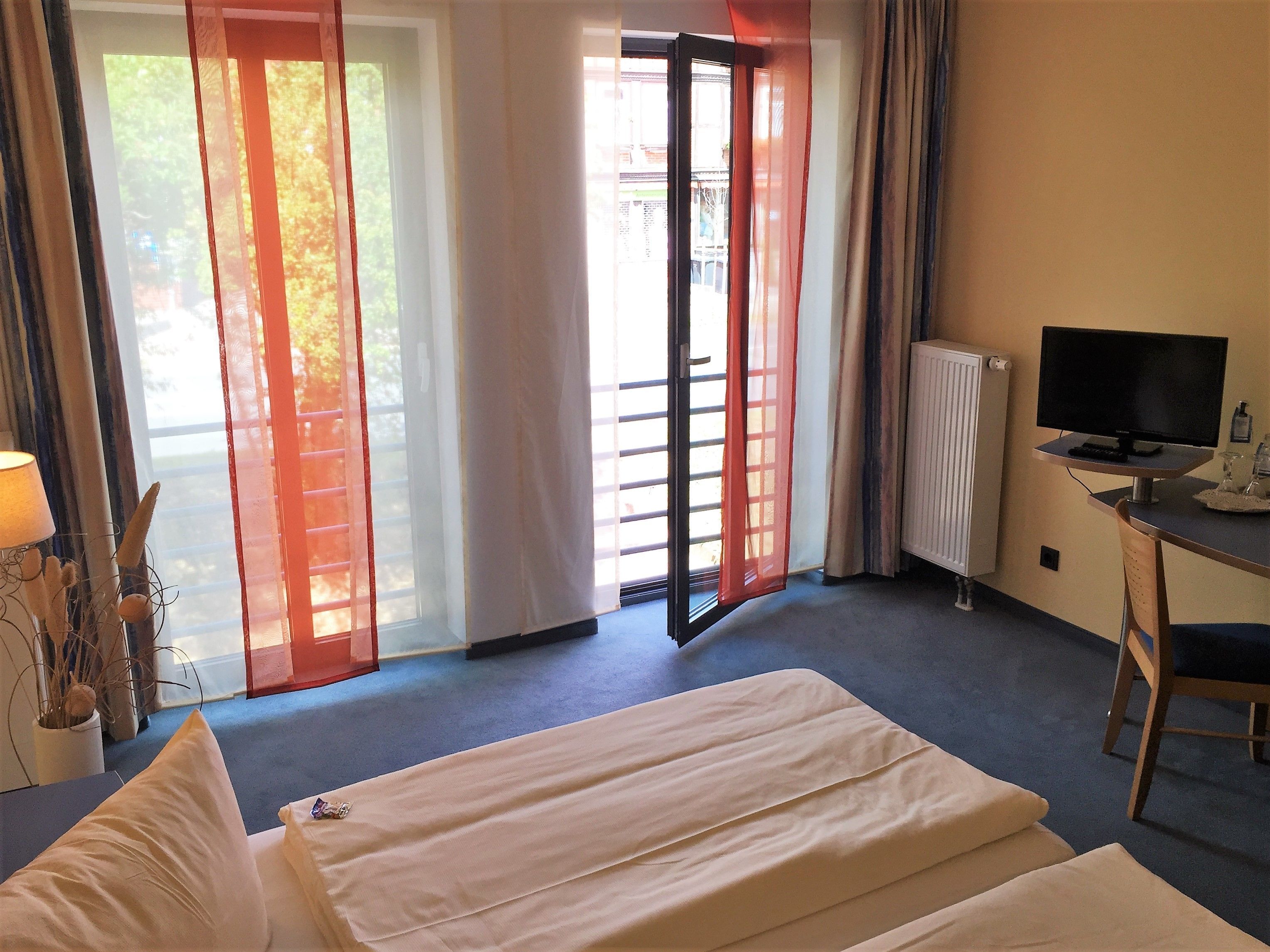 A hotel room with a bed, a desk with a computer, a radiator, and a view through a window with curtains.