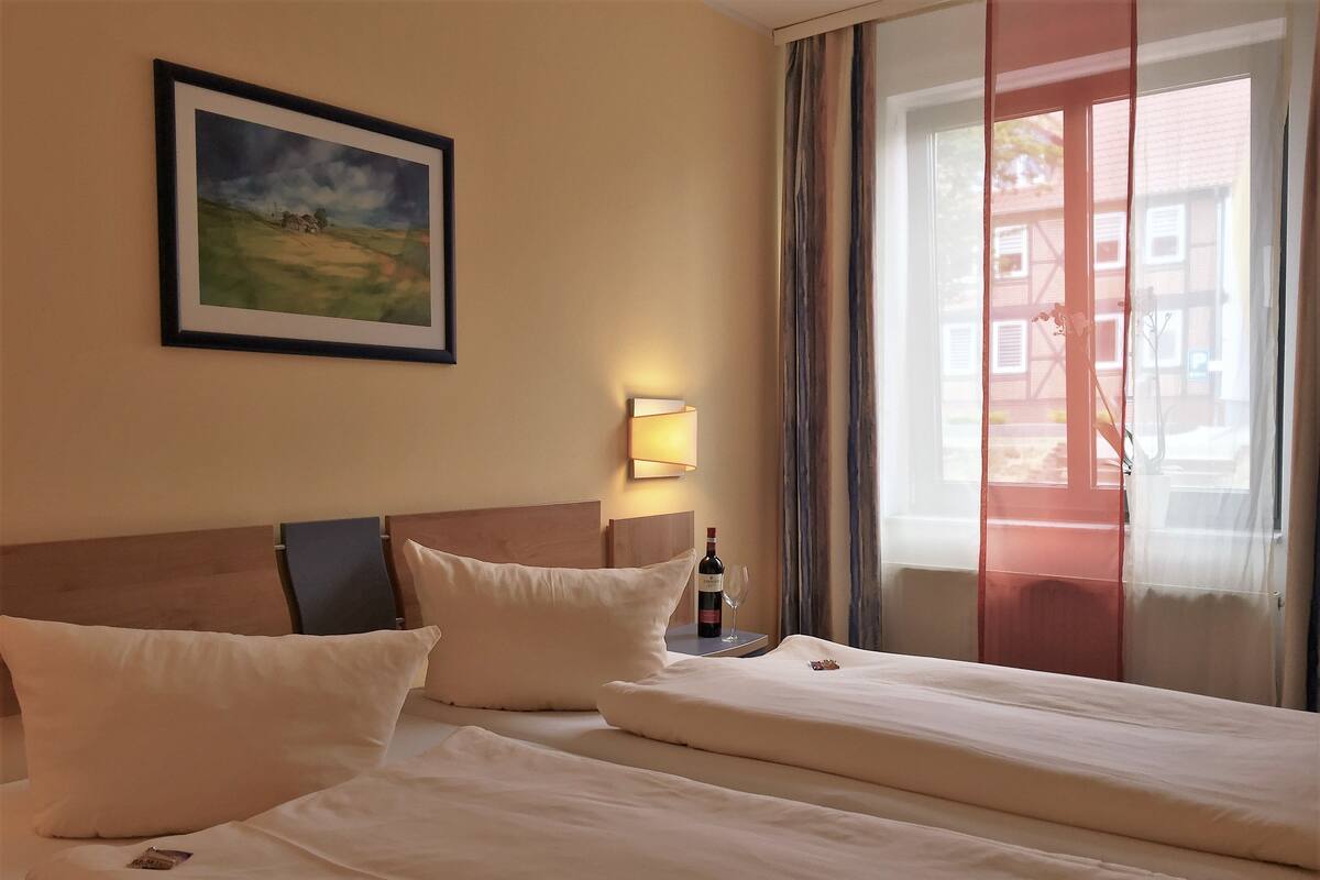 Une chambre d’hôtel avec deux lits, un tableau représentant un paysage encadré, une table de chevet avec une bouteille et un verre, et une fenêtre avec des rideaux.