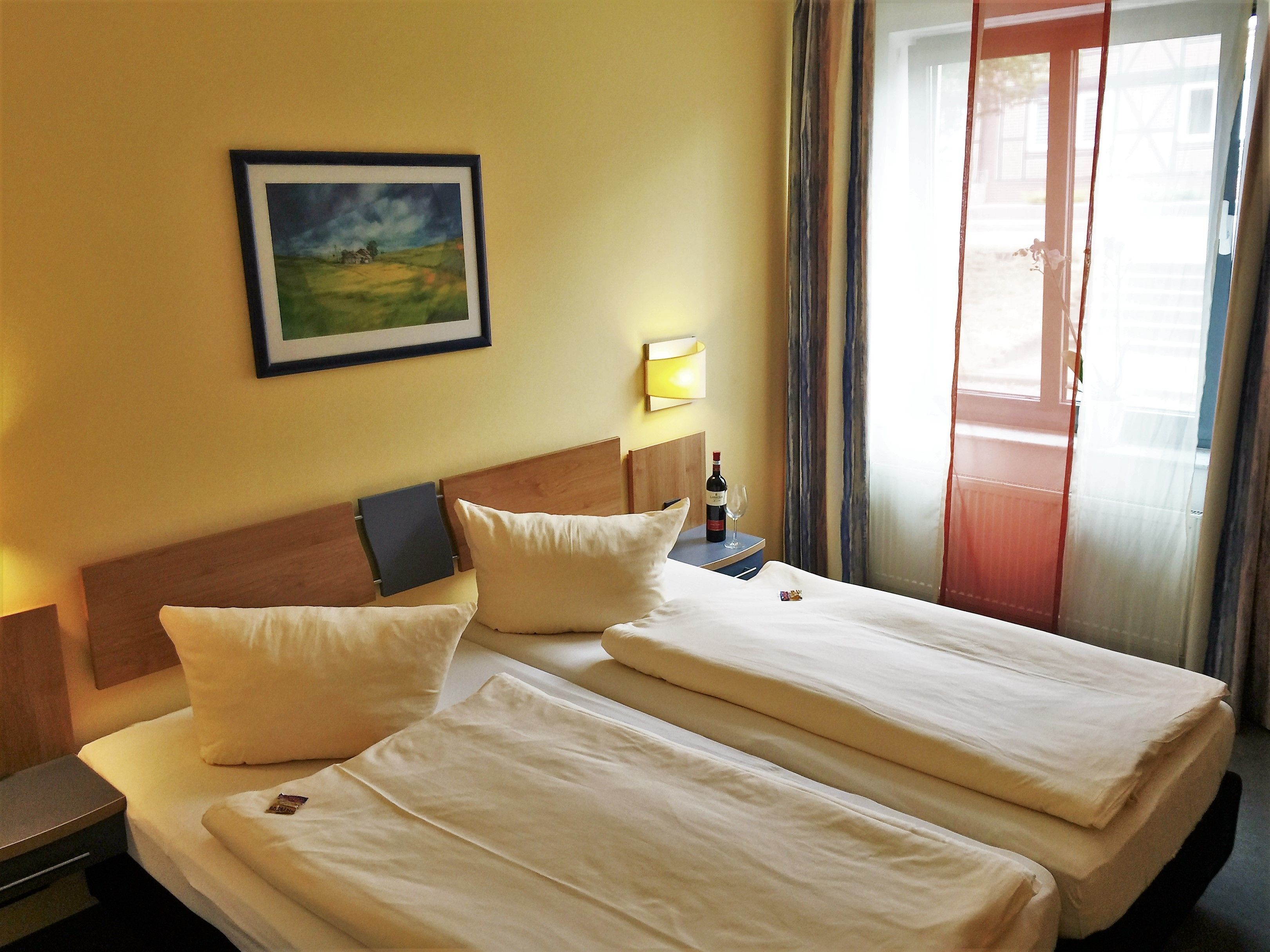 Habitación de hotel con dos camas, un cuadro en la pared, una botella de vino y una ventana con cortinas.