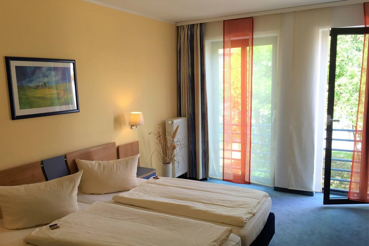 Une chambre d’hôtel avec deux lits, une table de chevet, une lampe, un tableau et une vue sur la verdure par la fenêtre.