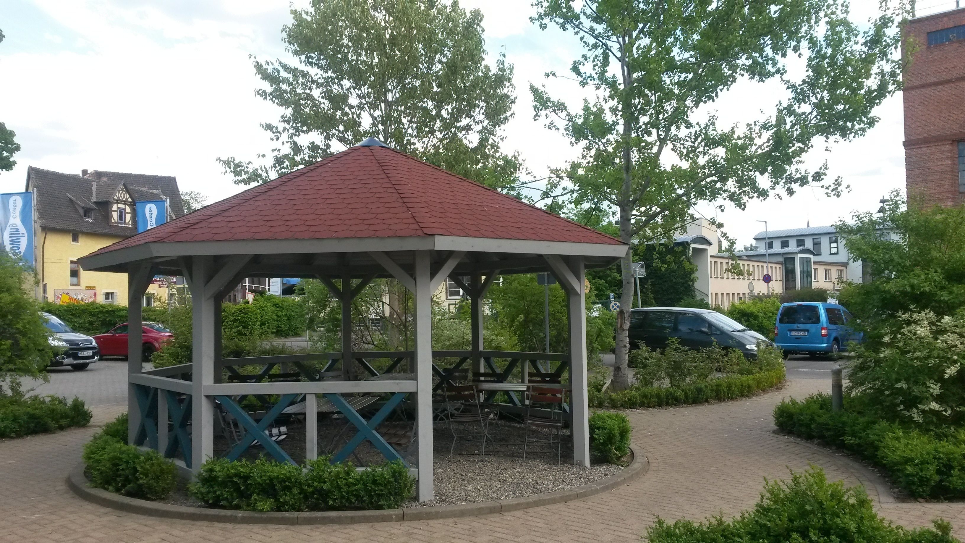 Um gazebo com telhado vermelho e estrutura branca, cercado por vegetação e um caminho pavimentado.