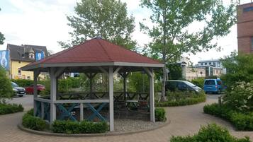 Un gazebo con tetto rosso e struttura bianca, circondato da vegetazione e un vialetto lastricato.
