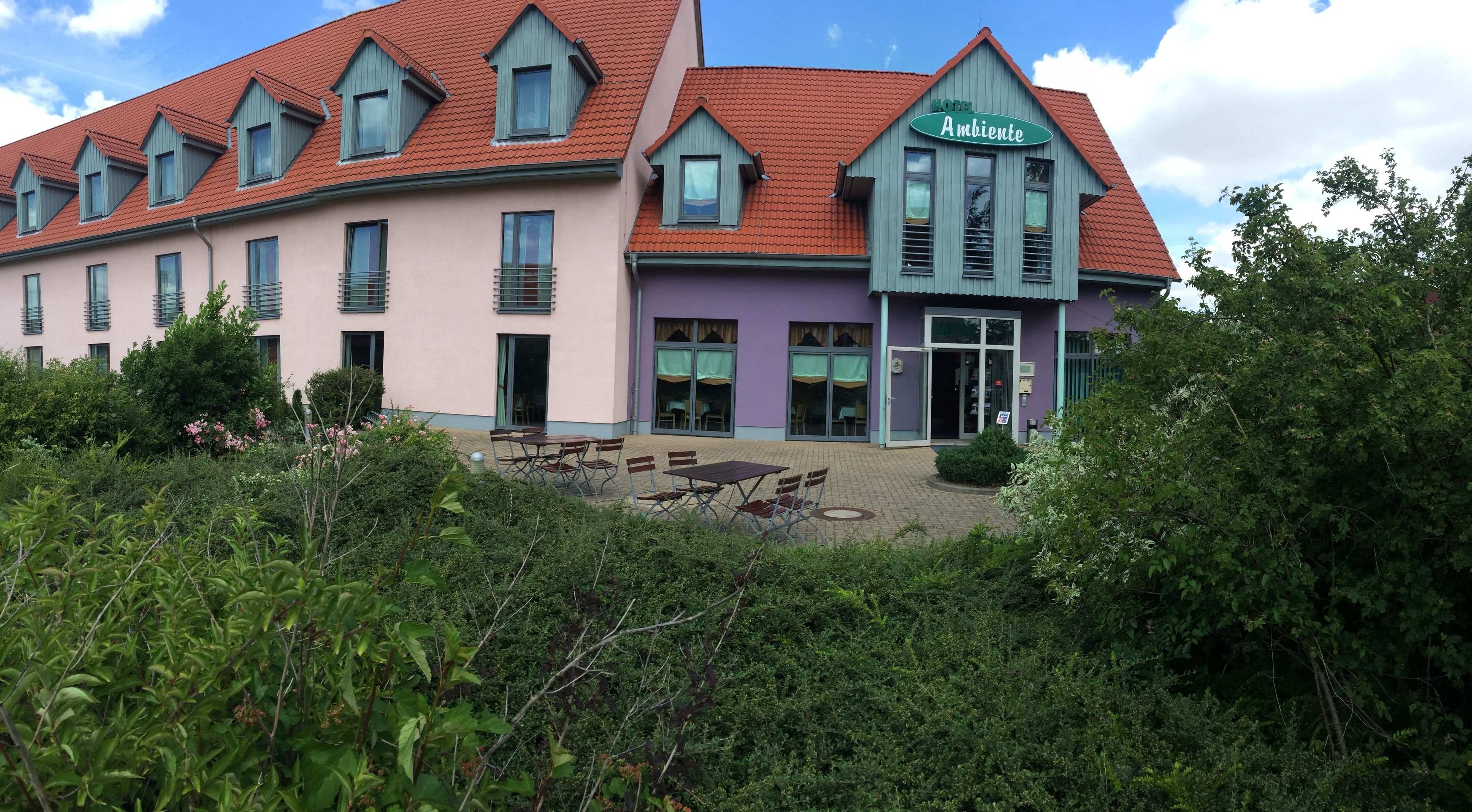 Ein Hotel mit rotem Ziegeldach, einem grünen Schild mit der Aufschrift "HOTEL Ambiente" und einer Terrasse mit Sitzgelegenheiten.