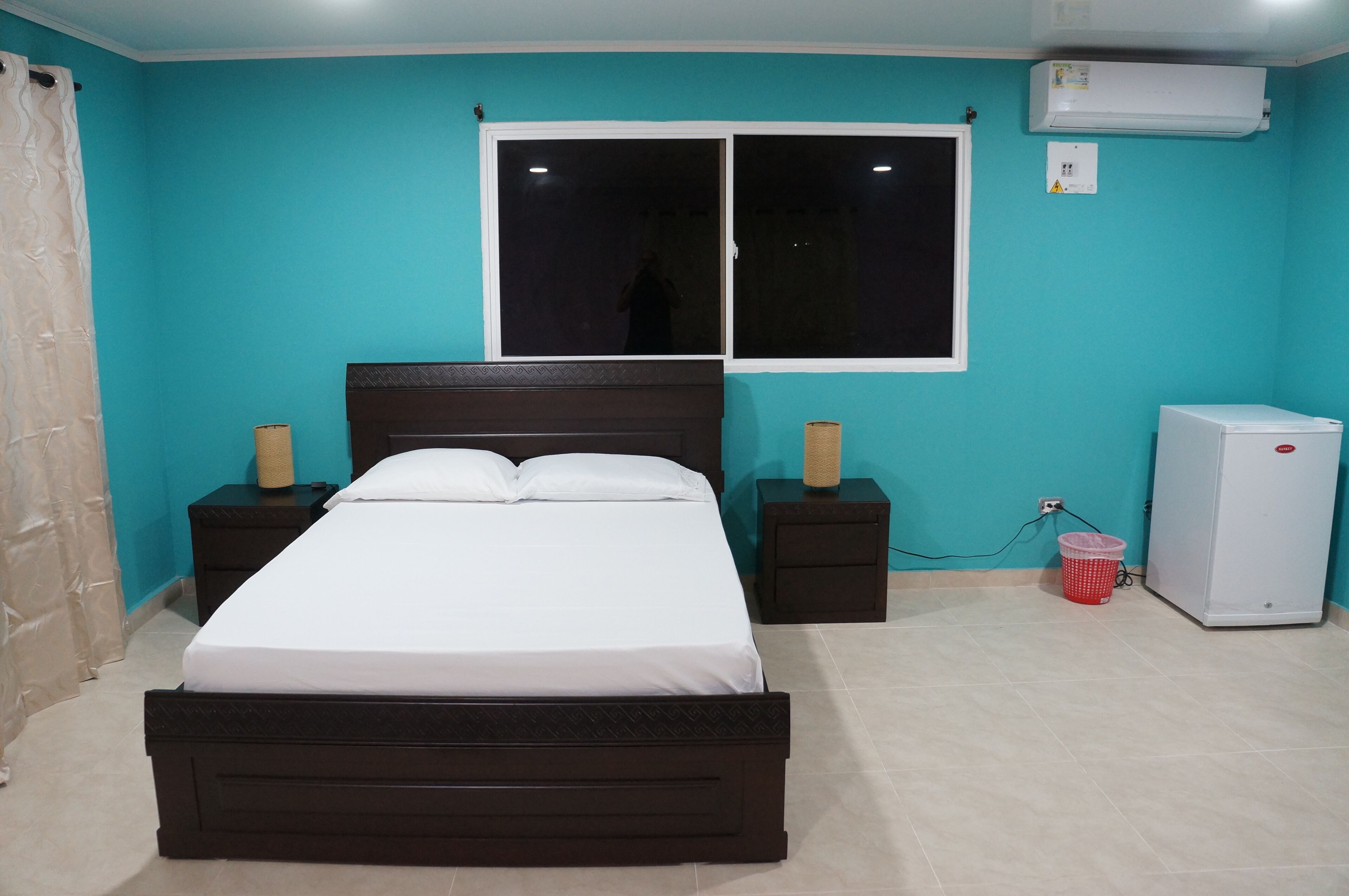 Un dormitorio con cama, mesitas de noche, ventana, aire acondicionado y refrigerador.