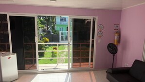 A room with a sliding glass door leading to a garden, a black sofa, a white refrigerator, and a standing fan.