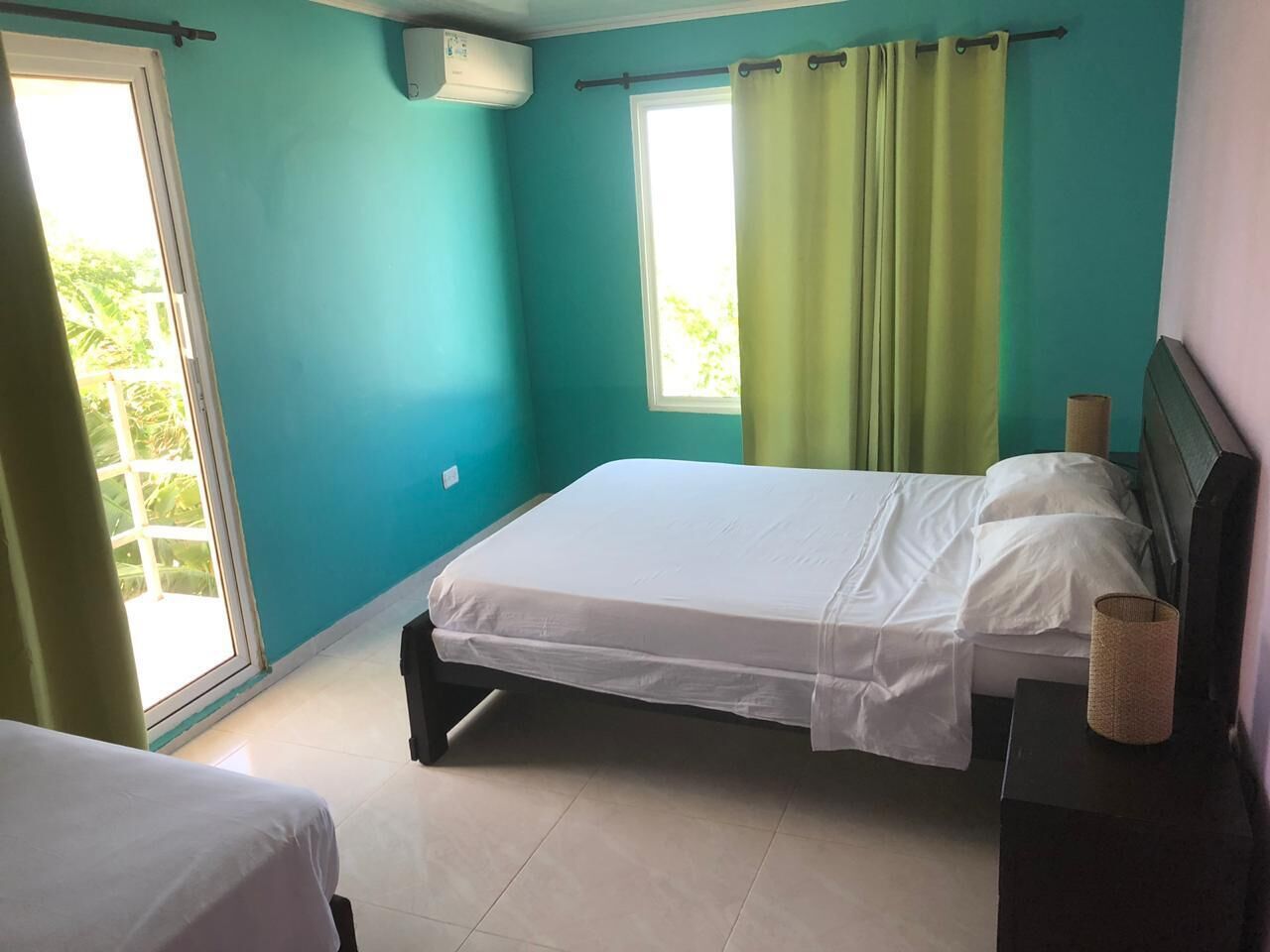 A bedroom with a bed, a bedside table, a window with curtains, and a view of greenery outside.