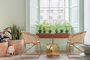 A room with two wicker chairs, a small table, and potted plants by a window with a view of buildings.