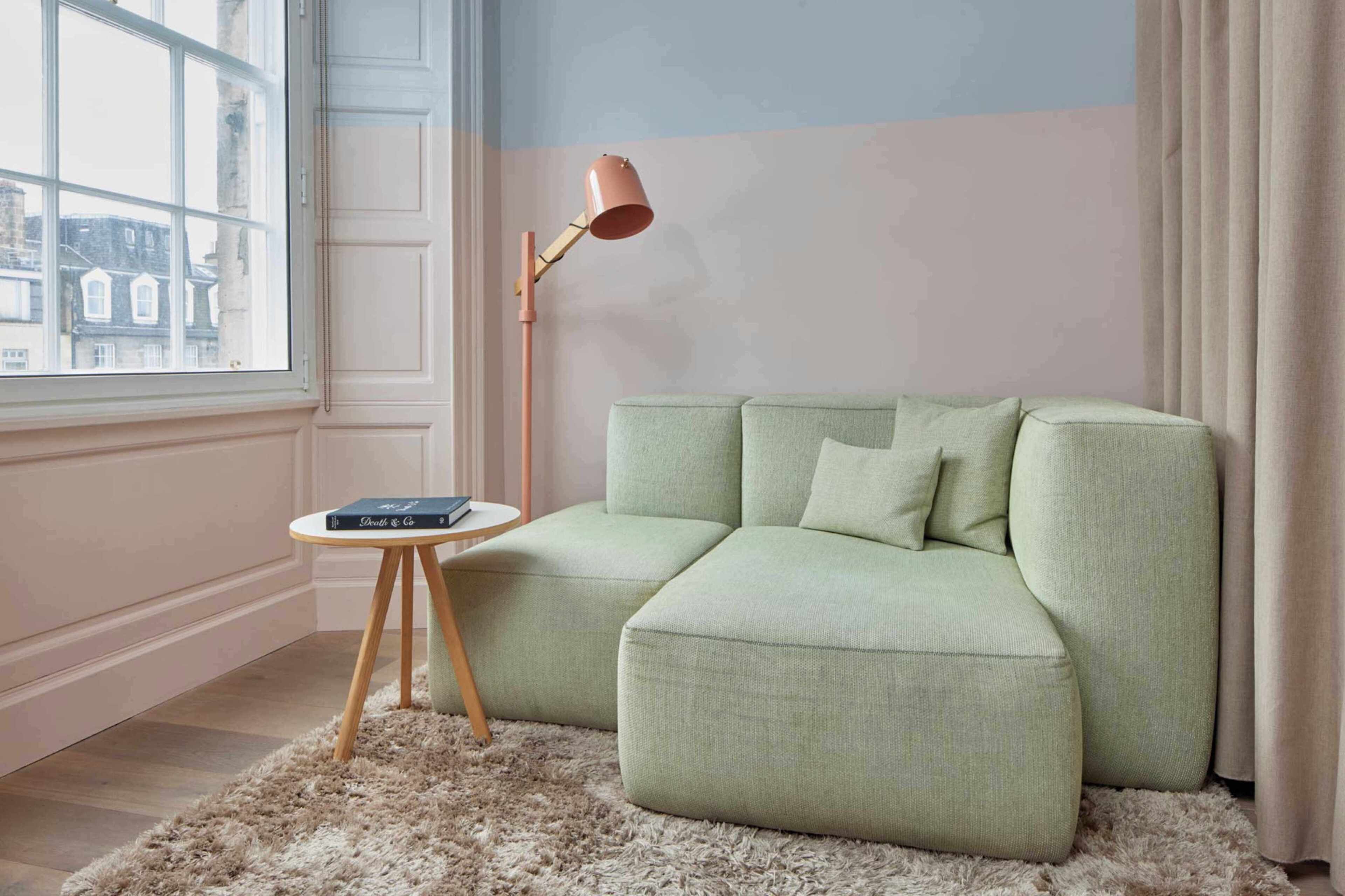 Ein Zimmer mit einem grünen Sofa, einem kleinen runden Holztisch mit Büchern, einer hohen Stehlampe und einem Fenster mit Blick auf Gebäude.