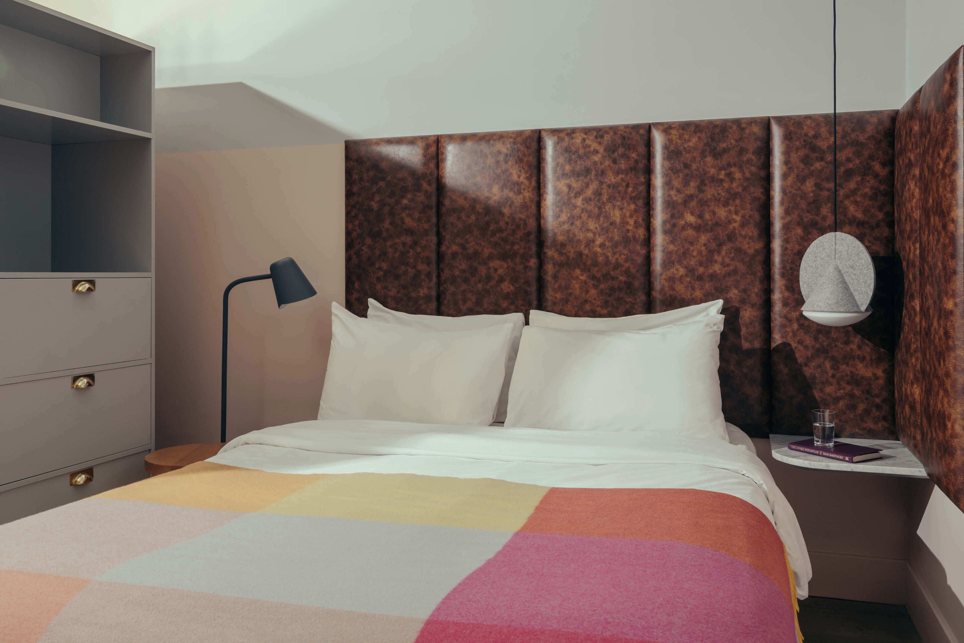Habitación de hotel con cama, mesita de noche con lámpara, una pequeña mesa con un libro y un vaso, y un armario con espejo.