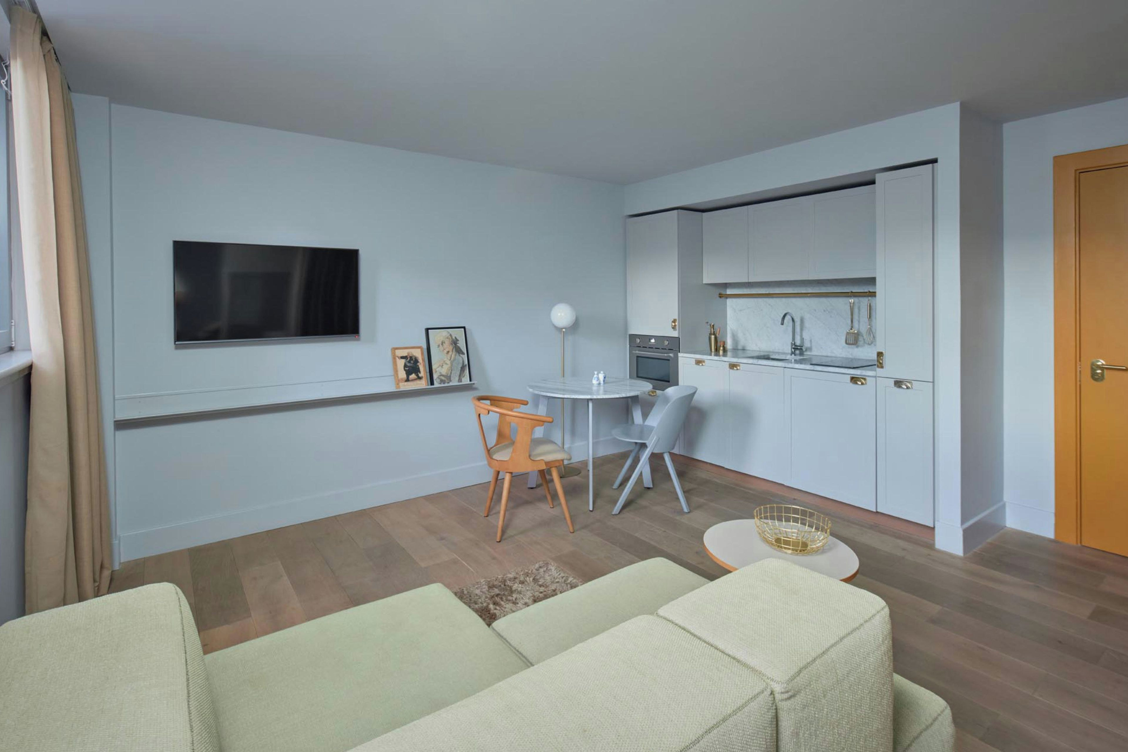 A modern living room with a sofa, a dining table with chairs, a kitchen area with cabinets, and a TV mounted on the wall.