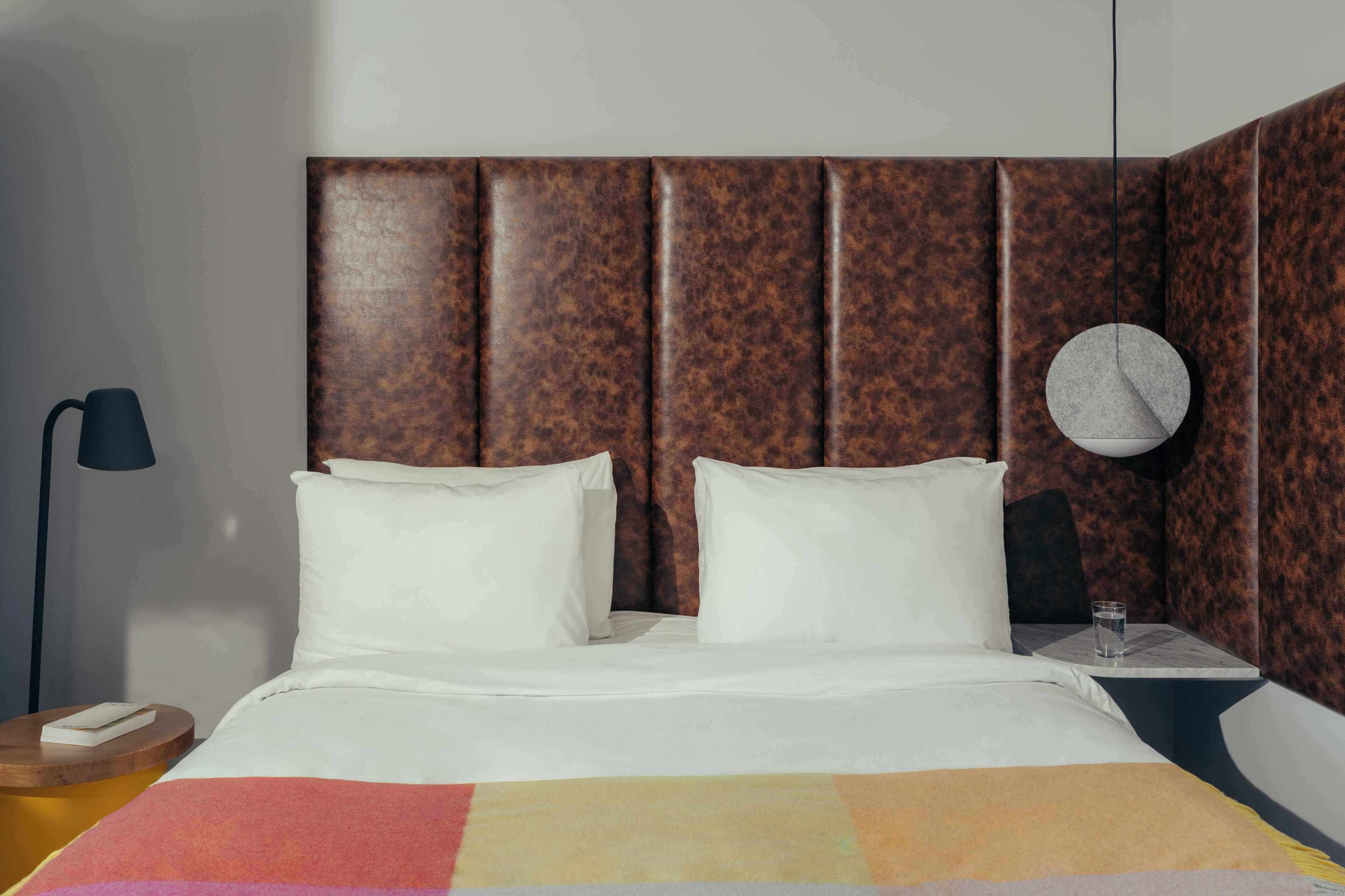 A hotel room with a bed, pillows, a bedside table with a lamp, and a wall clock.