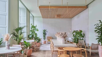 A spacious indoor area with multiple seating arrangements, including round tables and chairs, and several potted plants.