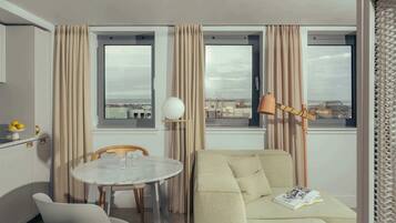 A hotel room with a dining table, a sofa, and a view of the cityscape through large windows.